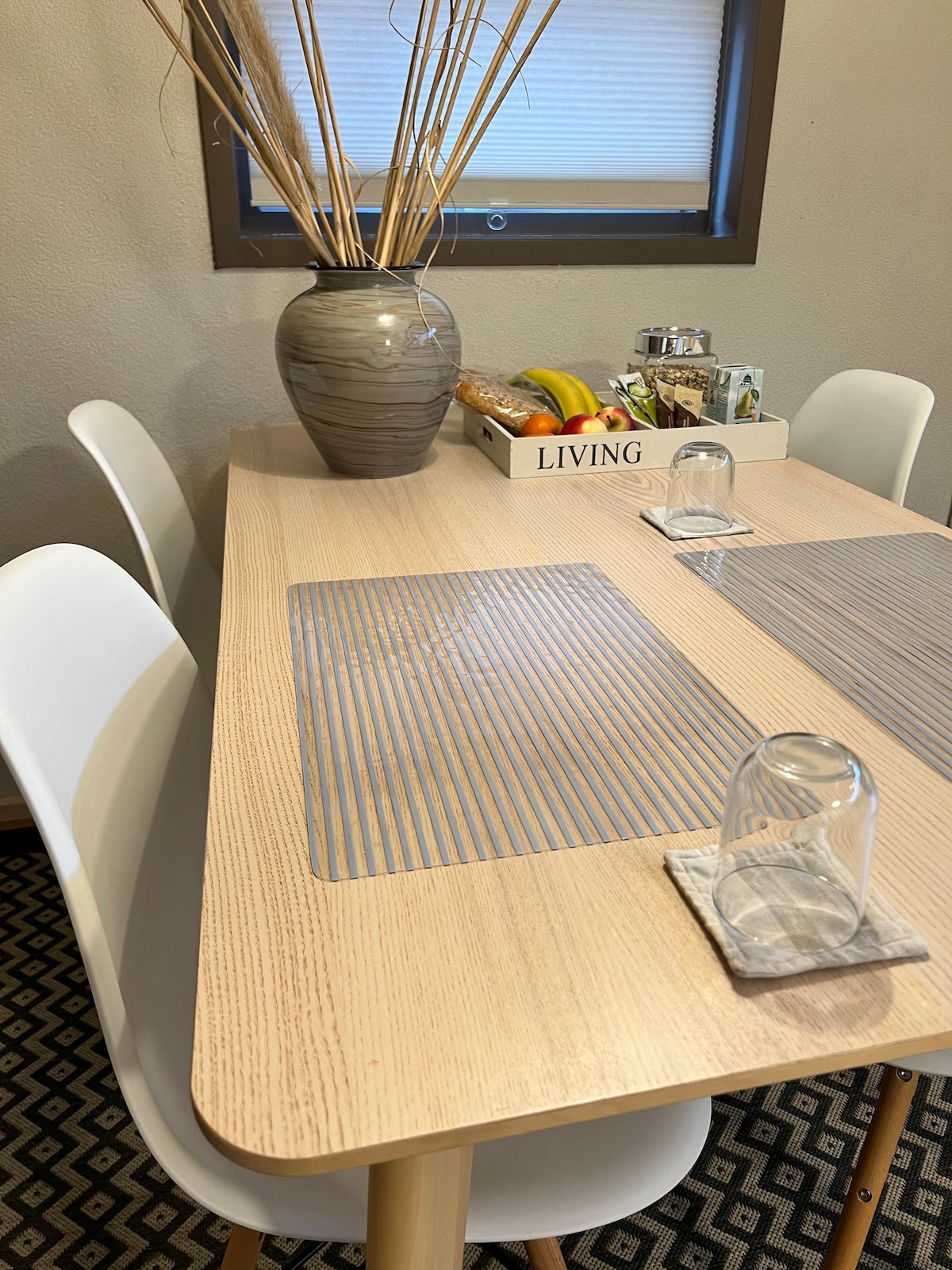 A dining table is set with four white chairs and placemats. A vase with decorative dried plants sits at the center, accompanied by a fruit bowl and a tray with assorted items. Two glasses rest on coasters, highlighting a functional and inviting dining area.