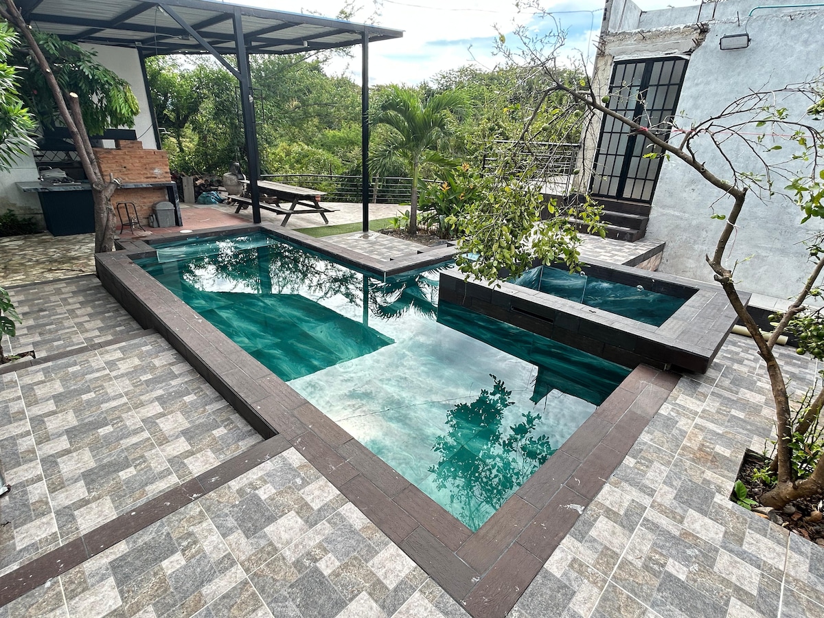 A private outdoor area features a unique multi-level pool with clear turquoise water, surrounded by a wooden deck. Lush greenery and stone pathways offer a natural feel, while a picnic table is positioned nearby, providing a space for relaxation and socializing.
