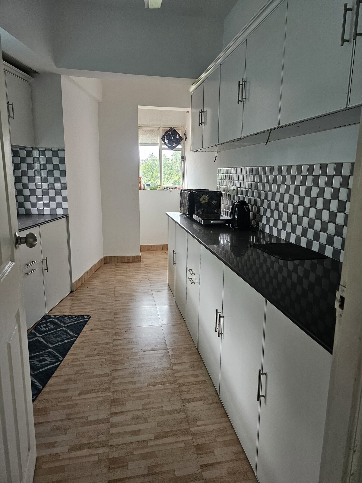 A modern kitchen area features white cabinetry paired with a black countertop. A small kettle and microwave are visible on the countertop. Light enters through a window, highlighting the checkerboard backsplash and tiled flooring. A blue mat is positioned on the floor.