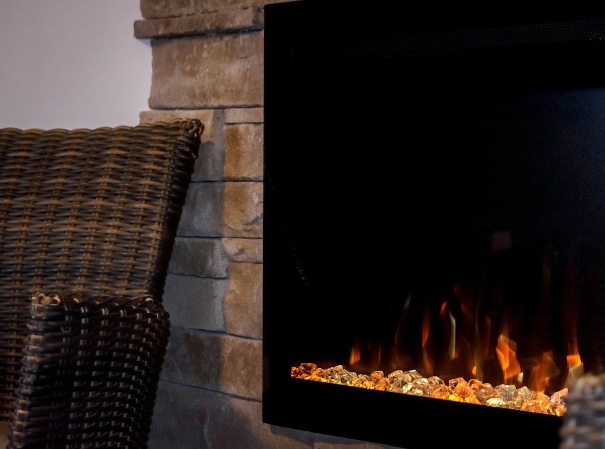 A stone fireplace with a contemporary design features flickering flames and decorative pebbles, providing a warm and inviting focal point. A woven chair is positioned nearby, creating a cozy seating area for relaxation.