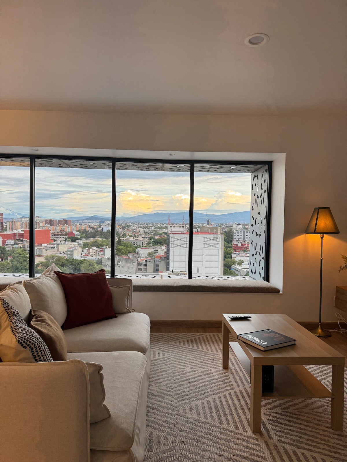 A bright living area features a comfortable sofa adorned with pillows, positioned next to a large window that offers expansive views of the city skyline. A small coffee table sits nearby, alongside a stylish floor lamp, enhancing the space's modern design.