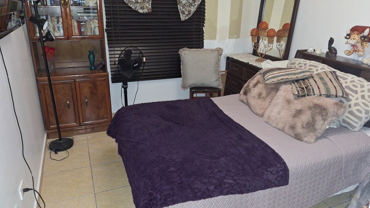 A cozy sleeping area features a spacious bed adorned with a textured purple blanket and various decorative pillows. A wooden dresser with decorative items is visible, along with a small fan and a floor lamp positioned nearby. Natural light filters through partially closed blinds.