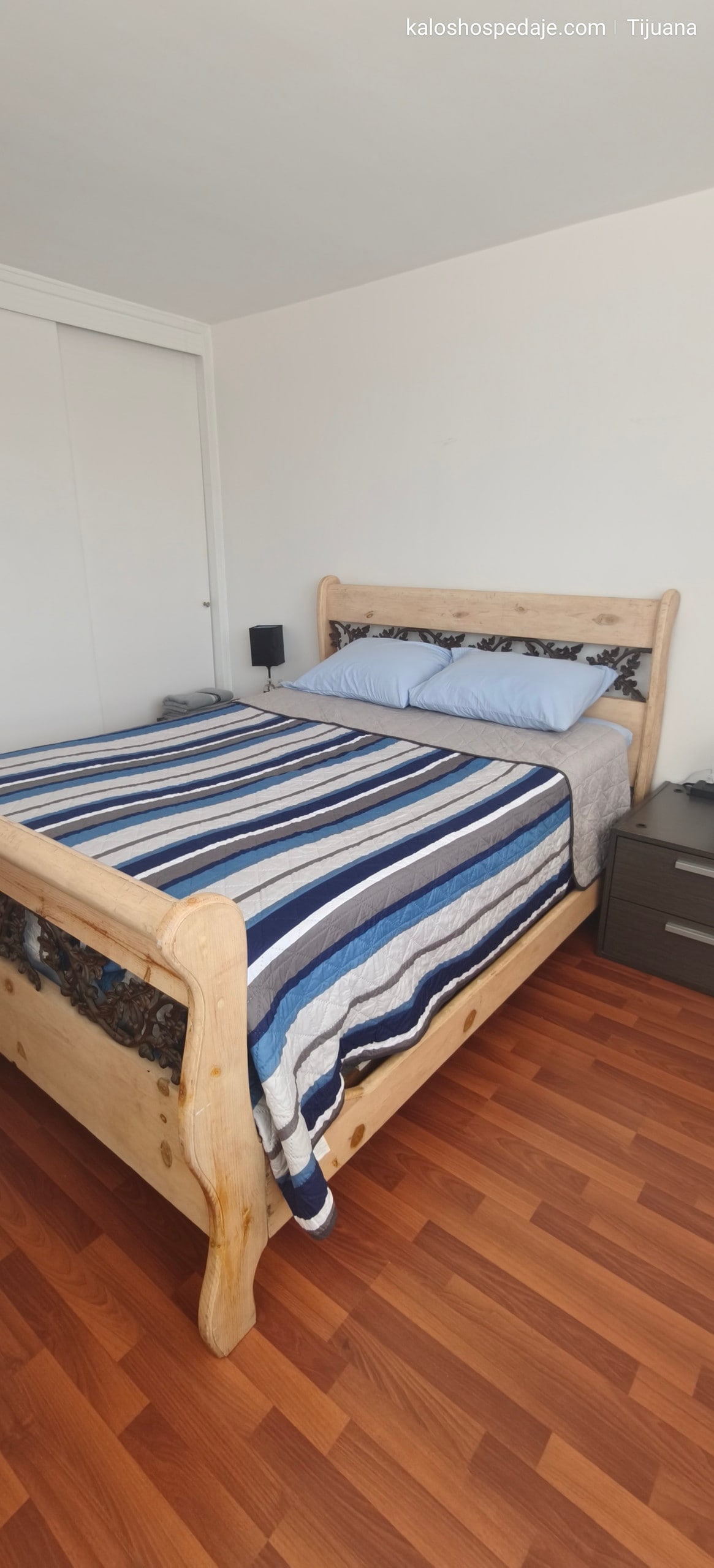 A spacious sleeping area features a wooden bed with a striped blanket in shades of blue and gray. Pillows are arranged at the headboard, and a nightstand with a lamp is positioned beside the bed. Light-colored walls enhance the room's brightness.