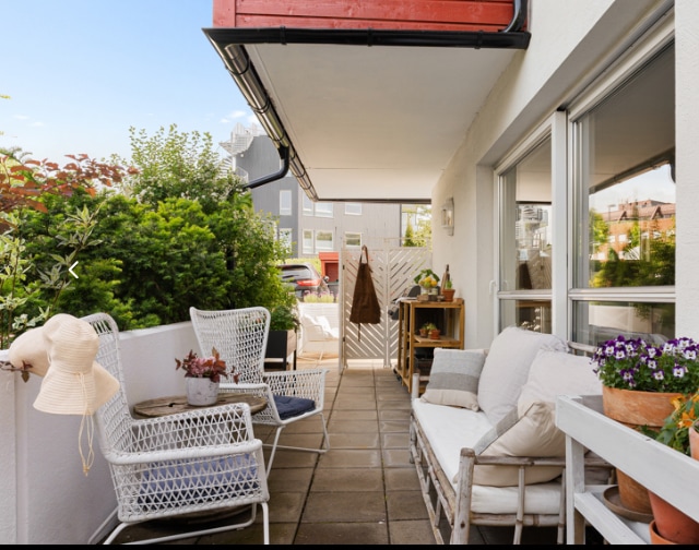 An outdoor patio area features a comfortable seating arrangement, including woven chairs and a lounge sofa. Greenery is present, adding a natural touch. A small table is adorned with potted plants, while bright sunlight illuminates the space.