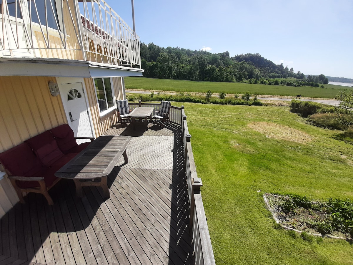 A spacious wooden deck is seen adorned with a large dining table and several outdoor chairs. Lush green fields and distant hills create a serene backdrop, while a clear sky provides abundant natural light to the area.