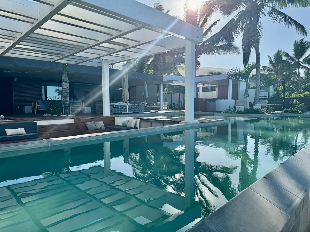 A serene outdoor area features a tranquil swimming pool reflecting the surrounding palm trees. Cozy lounge seating is positioned near the water, with sun loungers arranged for relaxation. The spacious patio is shaded by a modern pergola, promoting an inviting atmosphere.