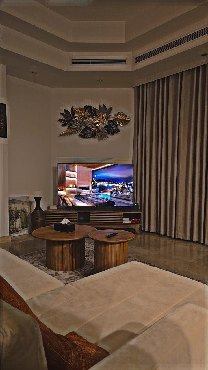 A modern lounge area is displayed, featuring a low wooden coffee table with a decorative item on top. A large television is mounted on the wall, accompanied by an elegant wooden media unit. Light-colored curtains frame the window, adding softness to the space.