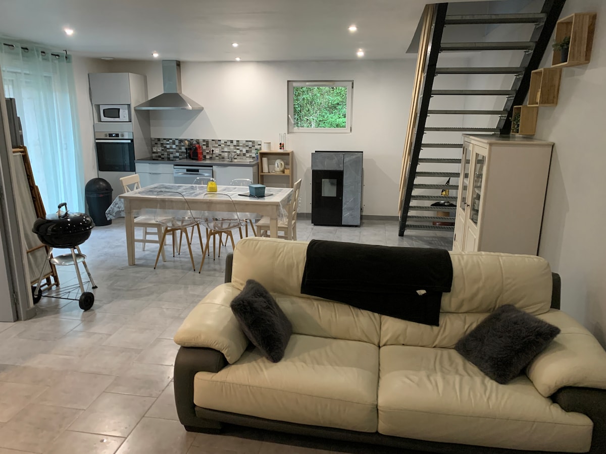 An open-plan living area features a spacious white sofa adorned with decorative pillows. A dining table with several chairs occupies the center, surrounded by a modern kitchen equipped with appliances and a stylish backsplash. A staircase leads to the upper level, while a barbecue grill stands nearby.