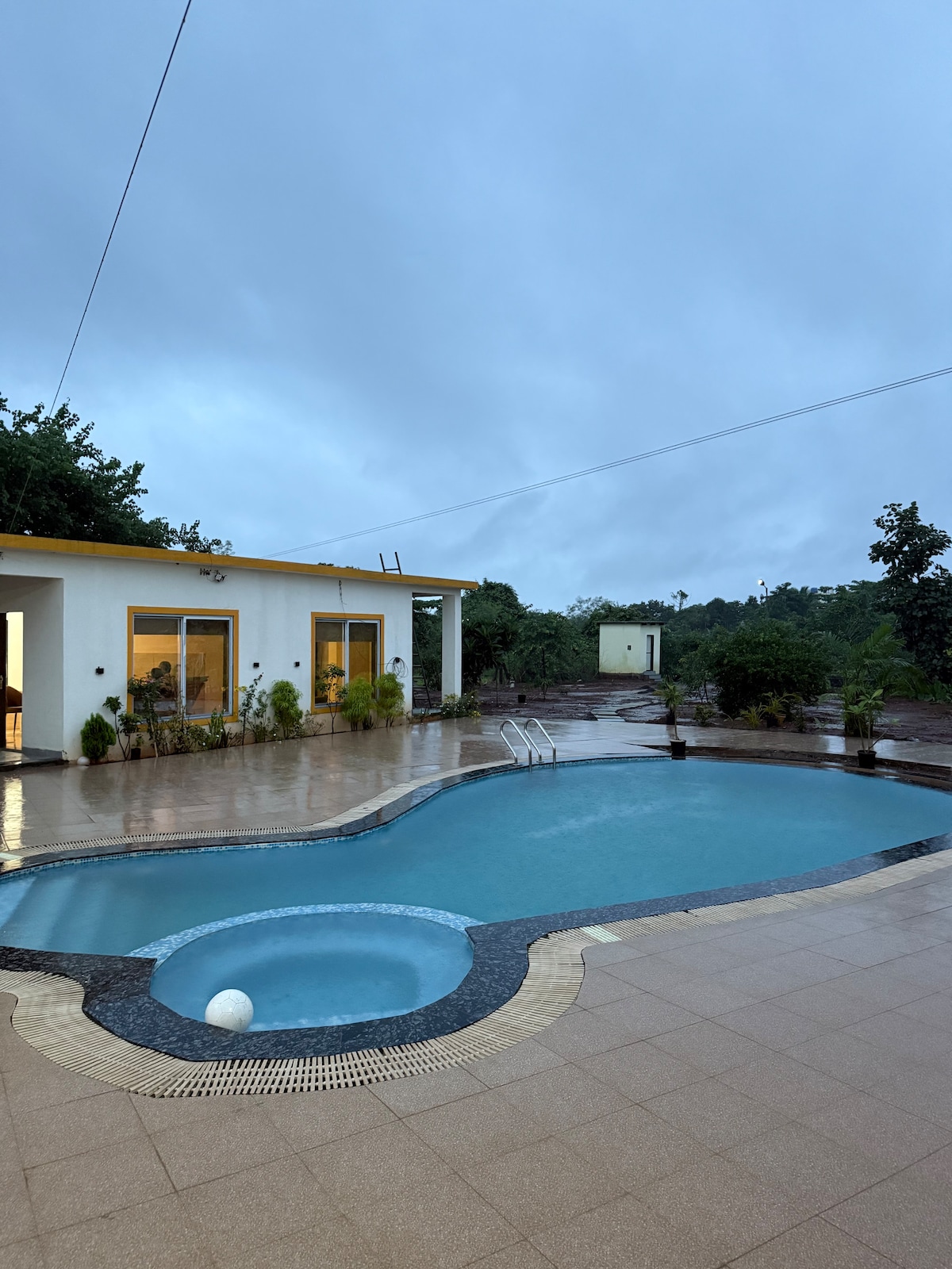 A refreshing swimming pool is framed by a spacious deck, featuring a whirlpool at one end. The pool is surrounded by lush greenery and a stone pathway, with light reflections on the water. The sky is overcast, indicating a serene atmosphere.