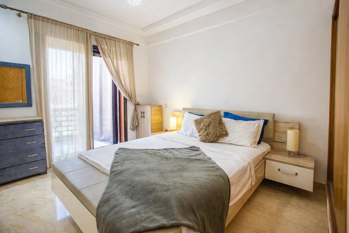 Модерна спална соба со двоен кревет и перничиња. Има лесна дрвена ноќна масичка со ламба и дрвен шкаф. Собата е светла и чиста и има поглед на мала кујничка која се гледа со отворот на вратата