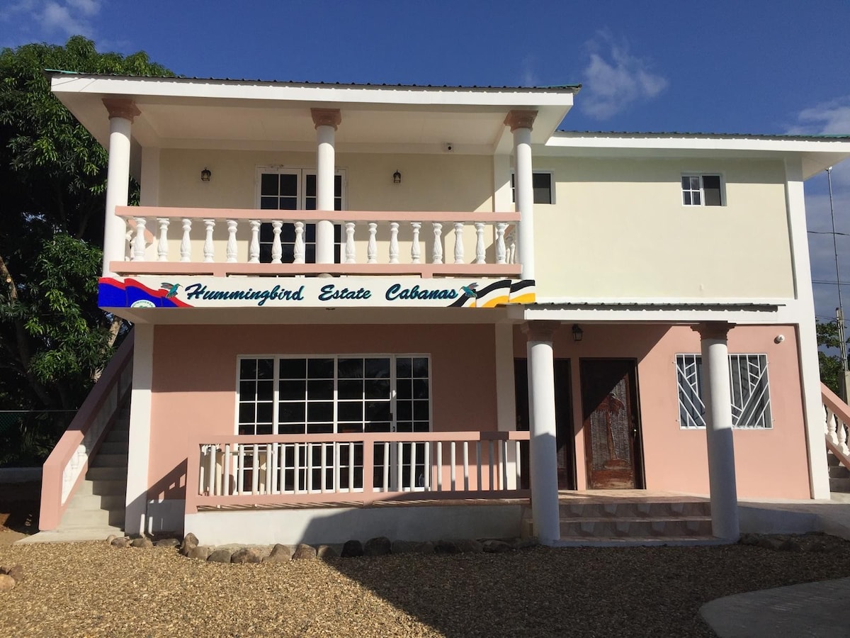 The exterior of Hummingbird Estates Cabanas is showcased, highlighting a two-story building with a welcoming entrance. A covered balcony with white railings overlooks a gravel pathway, and the colorful signage identifies the establishment. Lush greenery surrounds the property, contributing to a serene environment.