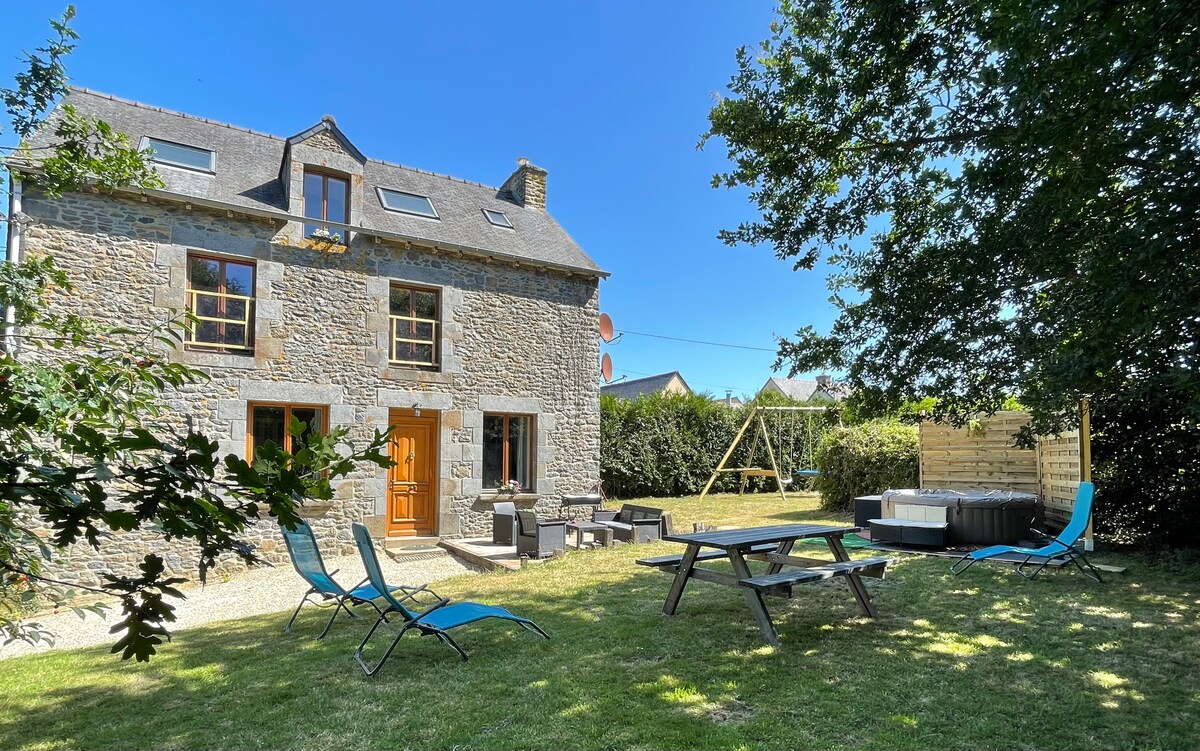 A serene outdoor space is visible, featuring a stone house with multiple windows and a wooden front door. Bright blue lounge chairs are arranged on the grass, alongside a picnic table. A Jacuzzi and swing set are positioned in a spacious garden framed by trees.