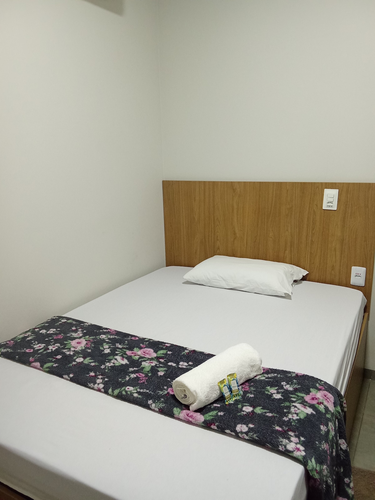 A simple sleeping area features a spacious bed with a light-colored sheet and a decorative floral throw draped across the foot. A single pillow rests at the head of the bed. The wooden headboard adds warmth to the minimalist setting.