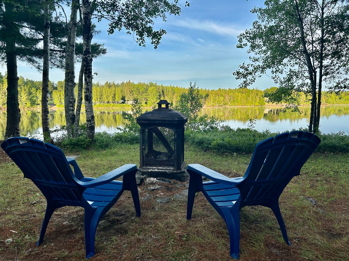 Two blue adirondack chairs are positioned near a fire pit, offering a view of the tranquil river and lush greenery. The calm water reflects the surrounding trees, creating a serene atmosphere for relaxation and gatherings.