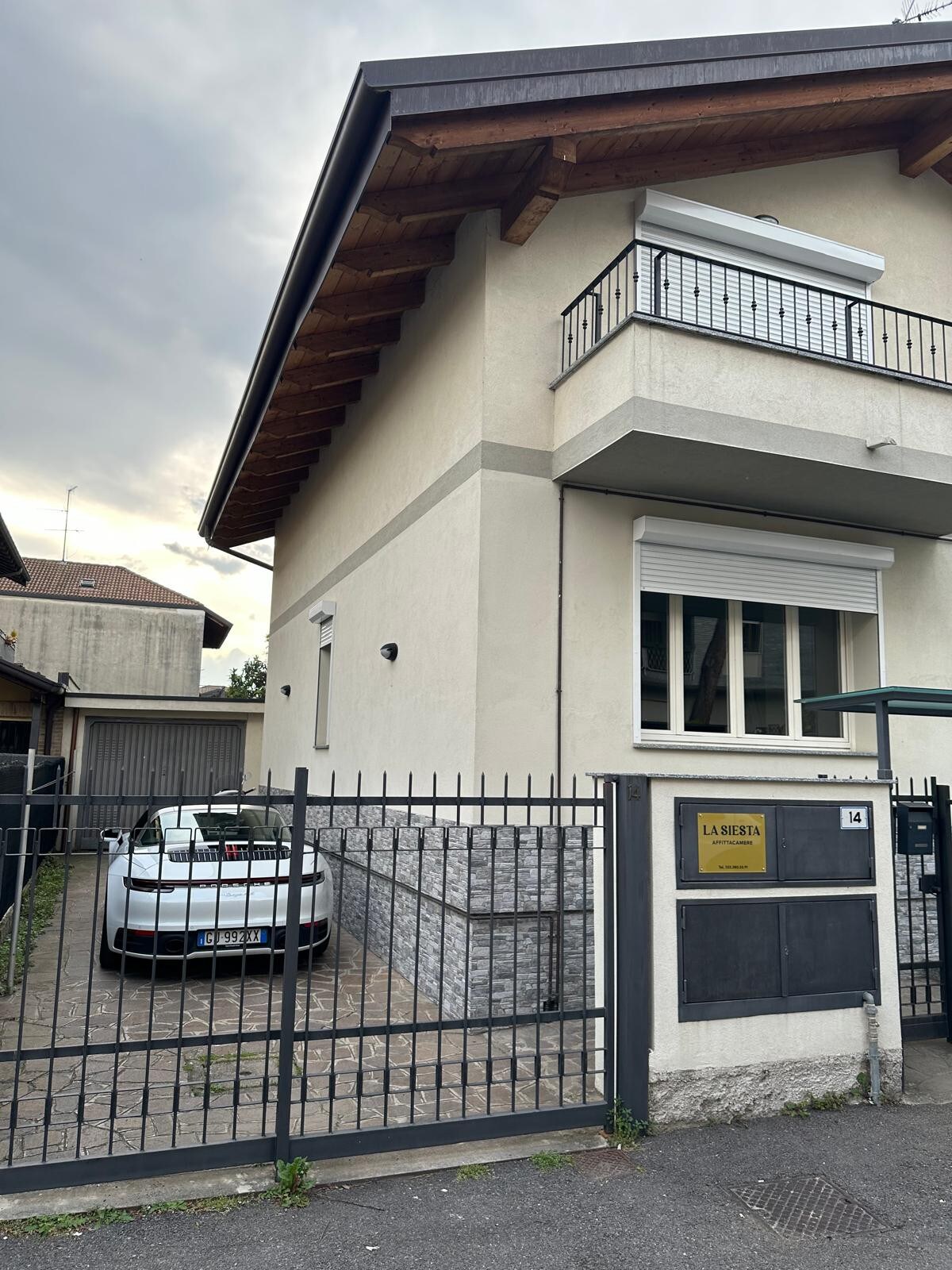A modern villa is depicted with a stone and stucco exterior, showcasing a fenced outdoor area. A parked car is visible in the driveway, and a covered entrance is positioned next to the window featuring horizontal blinds. The wooden roof overhang adds architectural detail.