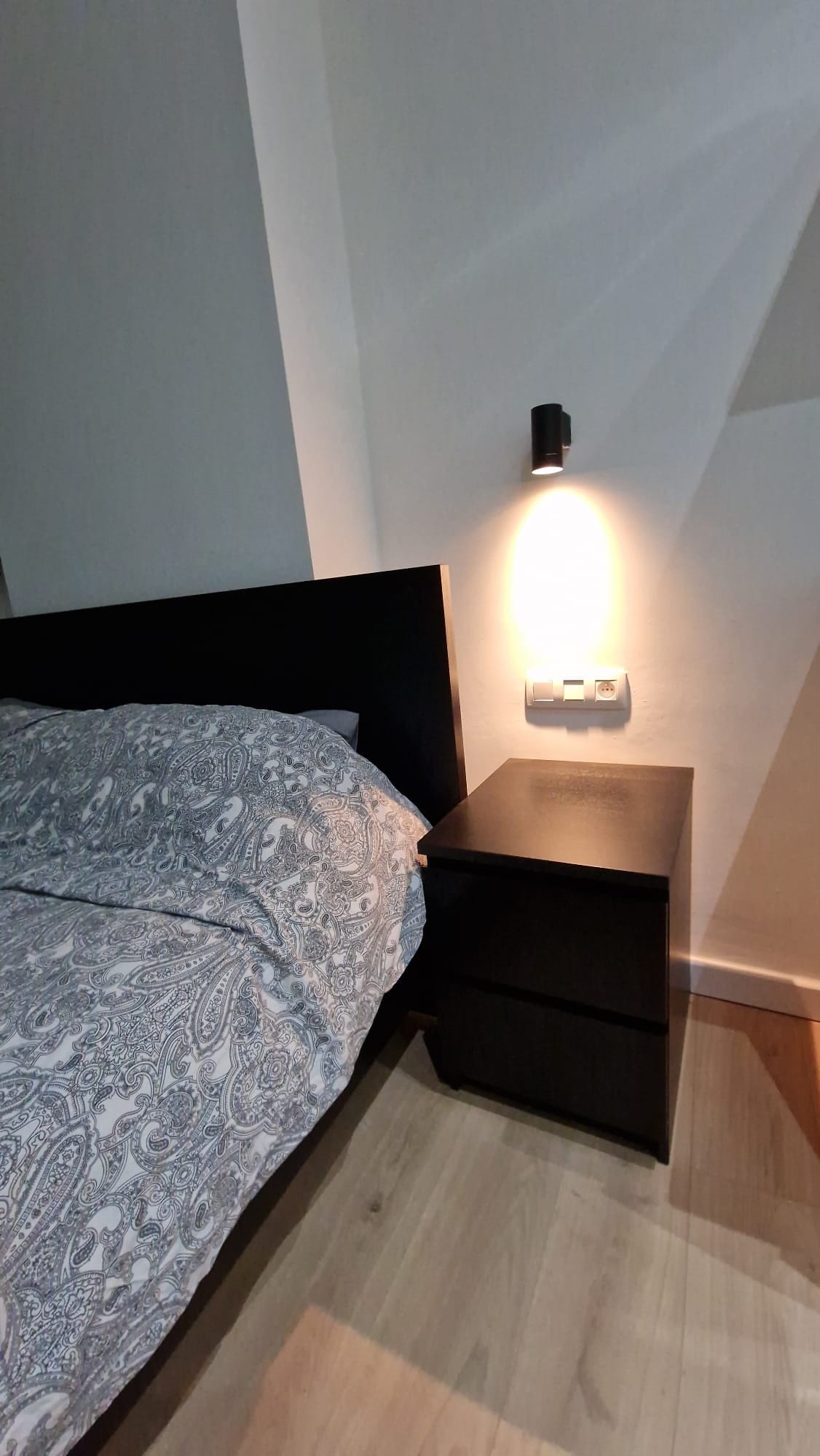 A cozy sleeping area features a spacious bed with a patterned duvet cover. A bedside table with a lamp is positioned beside the bed, offering warm light. The neutral wall and wooden floor create a calm and inviting atmosphere.