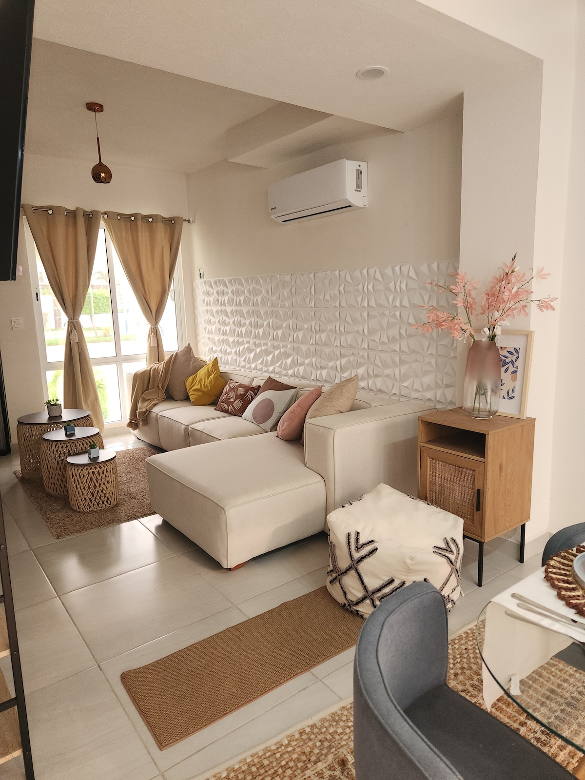 A comfortable living area features a light-colored sectional sofa adorned with patterned cushions. A decorative wall panel adds texture, while natural light streams in through large windows, dressed with sheer curtains. A small round table sits nearby, complemented by woven baskets and a neutral rug.