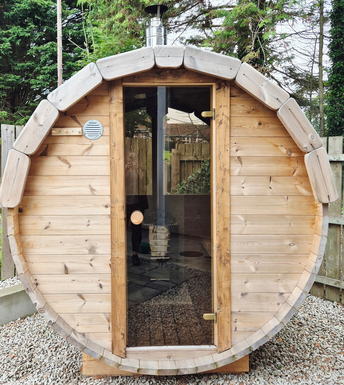 A wood-fired sauna features a circular wooden structure with a glass door. The exterior is constructed from natural wood planks, and the setting is surrounded by lush greenery. Pebbled ground complements the sauna, enhancing the outdoor relaxation space.