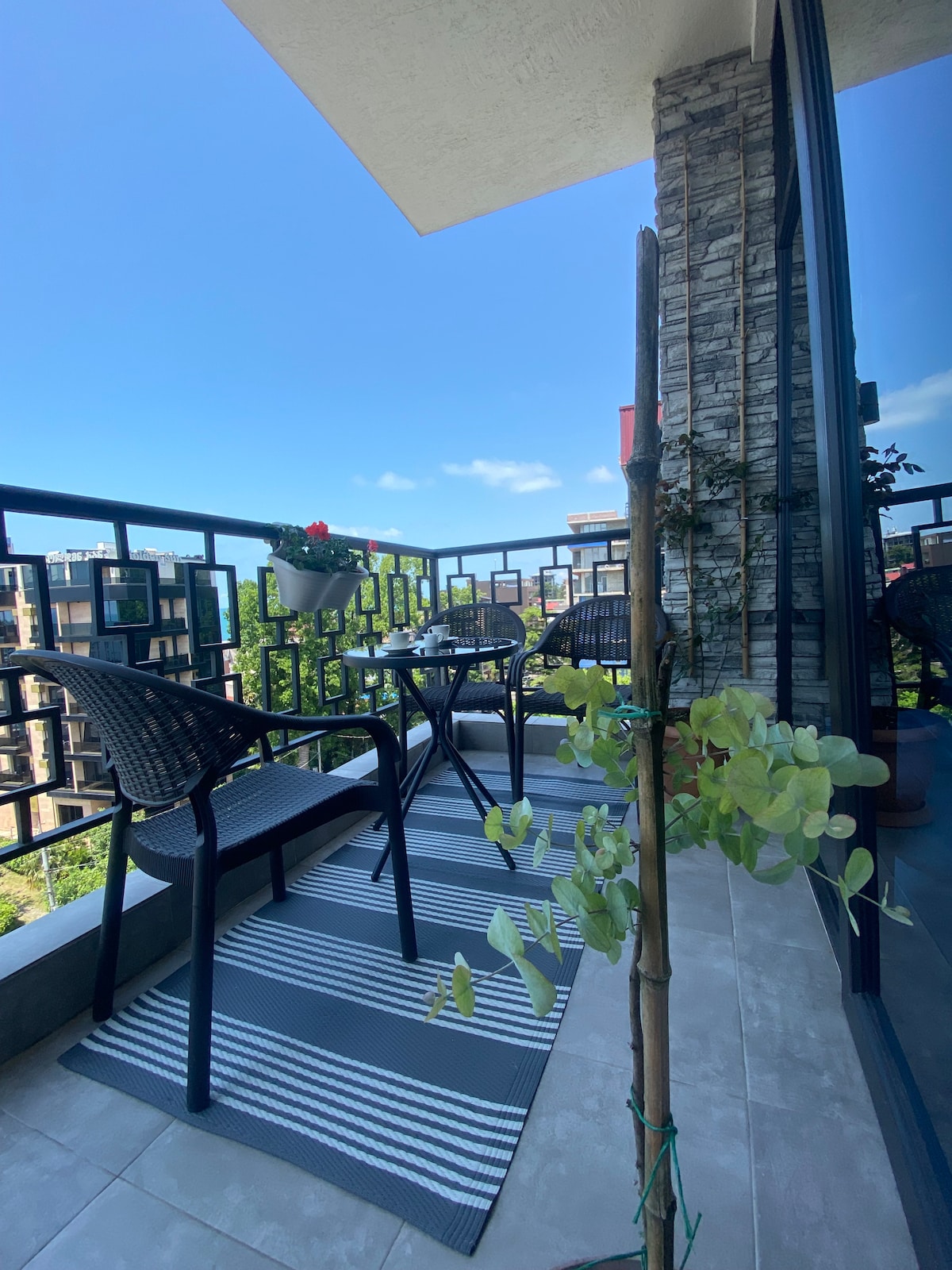 A balcony space features two black wicker chairs and a small table, positioned on a striped outdoor rug. Potted plants add greenery to the setting, while glass doors provide access to the interior. A clear blue sky enhances the inviting atmosphere.