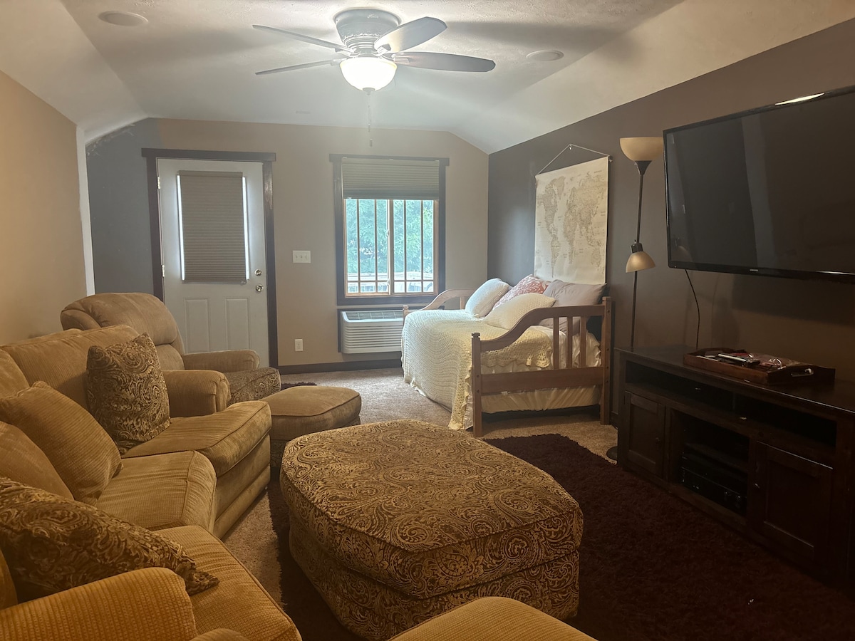 A cozy living area features a comfortable seating arrangement, including a plush sofa and armchairs. A large television is mounted on the wall opposite a daybed, inviting relaxation. Natural light filters through the window, while a soft area rug adds warmth to the space.