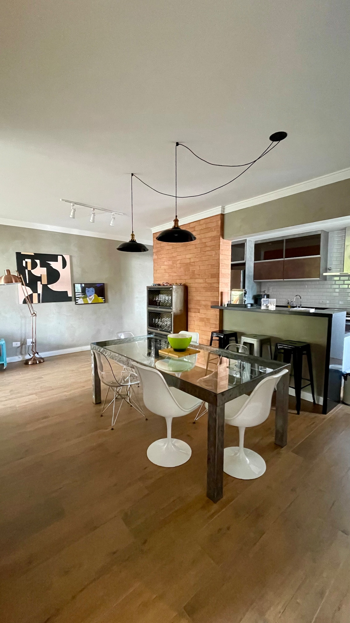 A dining area features a modern glass table surrounded by white chairs. Above, two pendant lights provide illumination. A decorative bowl sits at the center of the table, with a brick accent wall visible in the background. A well-equipped kitchen can be seen through an open space.