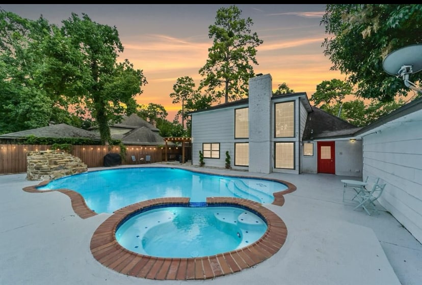 A spacious backyard is highlighted by a large swimming pool with an integrated hot tub. The pool area is surrounded by white concrete and features lush greenery in the background. The house showcases a contemporary design, with large windows reflecting the evening sky.