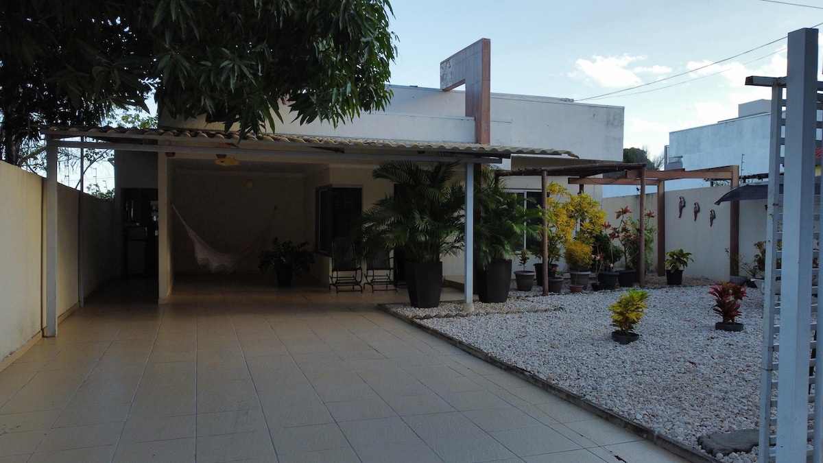 The outdoor area features a spacious patio with tiled flooring and a covered dining space. Various potted plants add greenery, while a hammock is seen nearby, offering a leisurely spot. The setting is enclosed by walls, creating a tranquil environment.