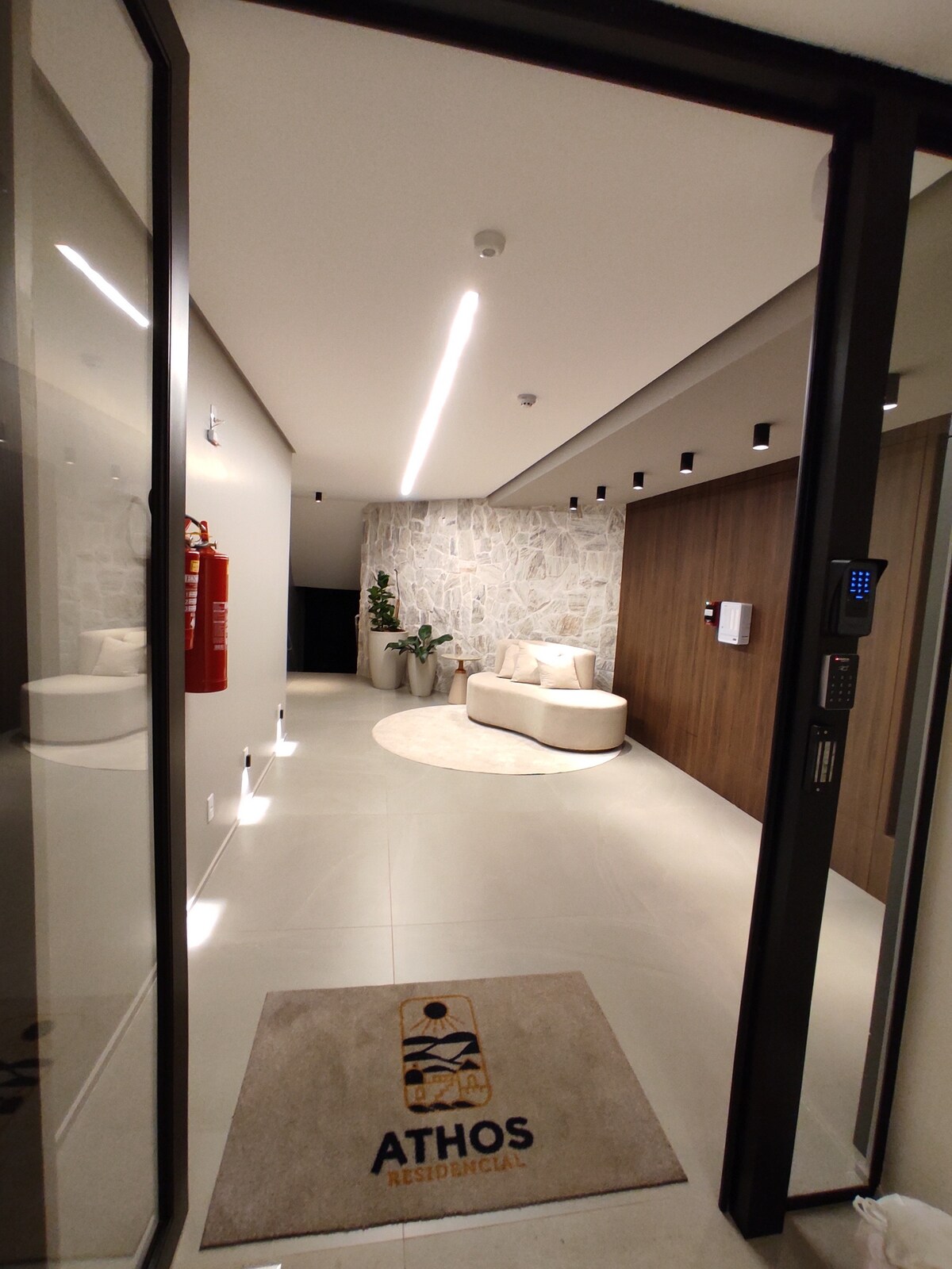 A welcoming entrance features a circular area rug on tiled flooring, with a modern seating arrangement against a textured stone wall. Soft lighting illuminates the space, highlighting contemporary design elements. Safety equipment is visible for convenience and security.