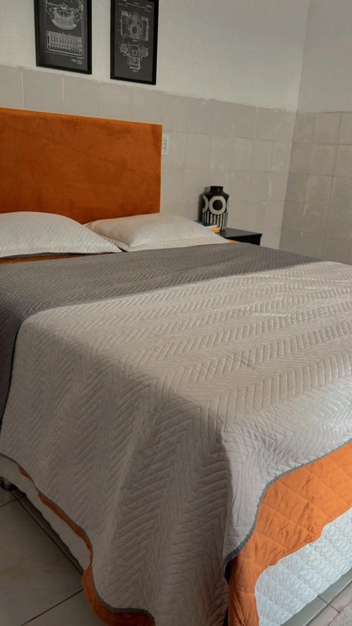 A spacious bed is covered with a textured gray and orange quilt. Two pillows rest against an orange headboard. A small bedside lamp is visible on a dark nightstand, and framed artwork adorns the wall above the bed, creating a calm ambiance.