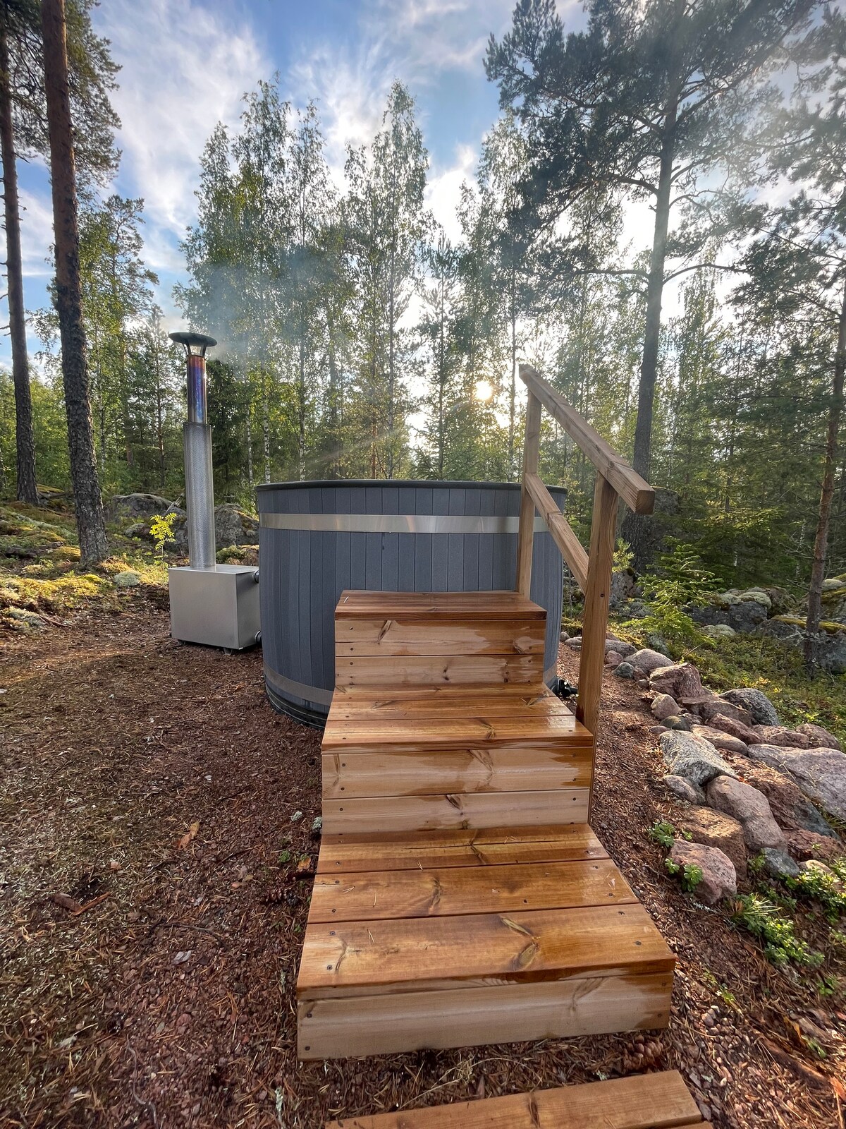 A wooden staircase leads up to a circular hot tub, positioned among trees. Steam rises from the tub, and natural light filters through the foliage, creating a serene setting. A heating unit stands nearby, enhancing the inviting atmosphere for relaxation.