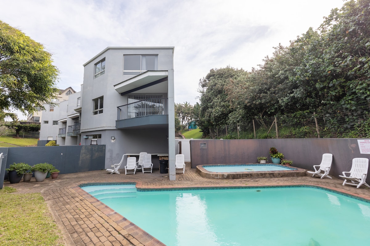 A well-maintained pool area features a light blue swimming pool surrounded by a brick patio. Several white loungers are arranged neatly around the pool. A hot tub is positioned nearby, with greenery and hedges providing a natural backdrop.