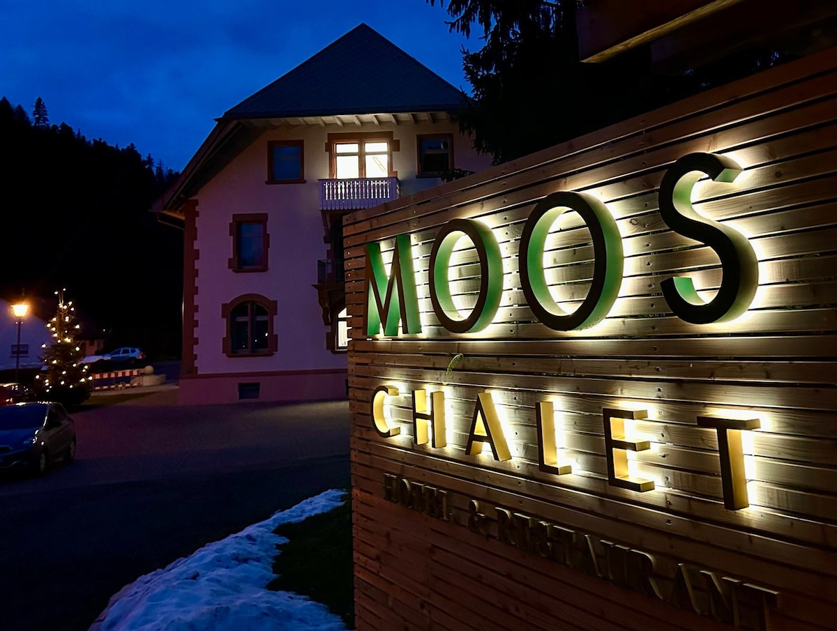 Chalet Moos is featured at night with illuminated signage prominently displayed against a wooden backdrop. The building's exterior is softly lit, revealing architectural details, while the surrounding area shows hints of snow and festive lights accentuating the inviting ambiance.