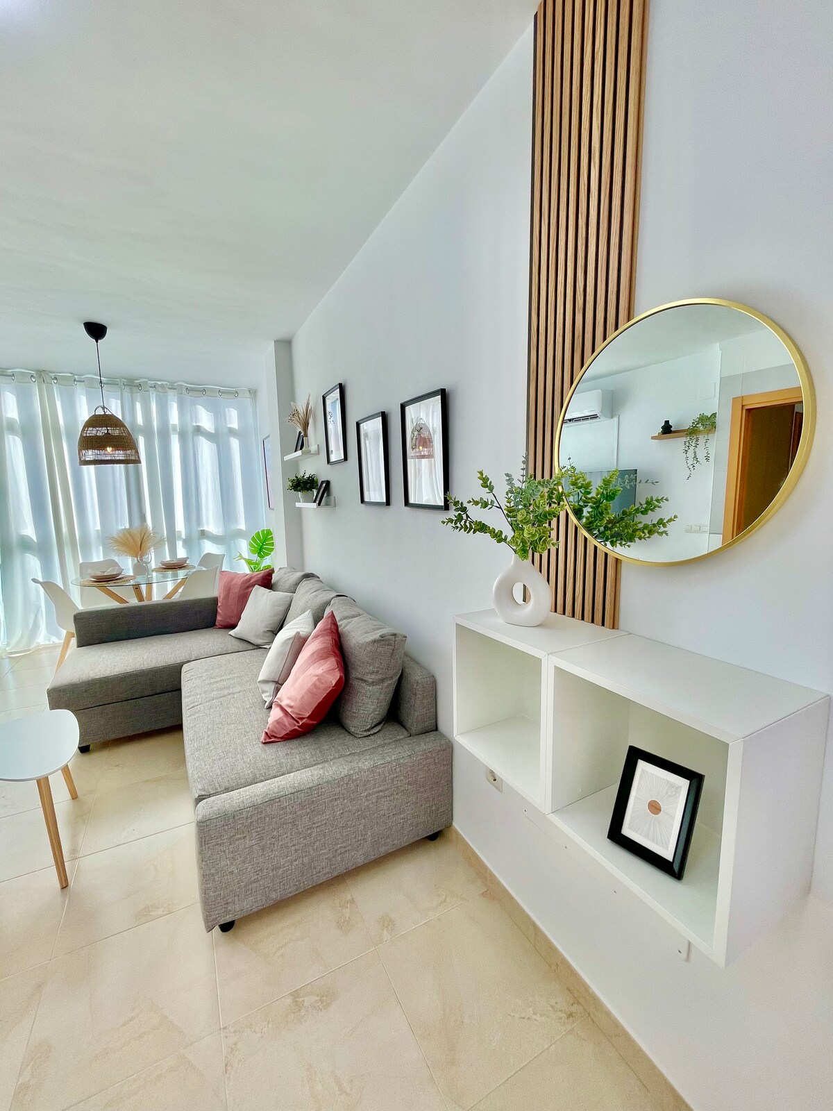 The living area features a modern grey sofa adorned with accent pillows, a circular mirror, and minimalist wall art. A light-filled dining space is visible through sheer curtains, with a round table and four chairs positioned nearby. A decorative shelf holds a small plant and framed artwork.