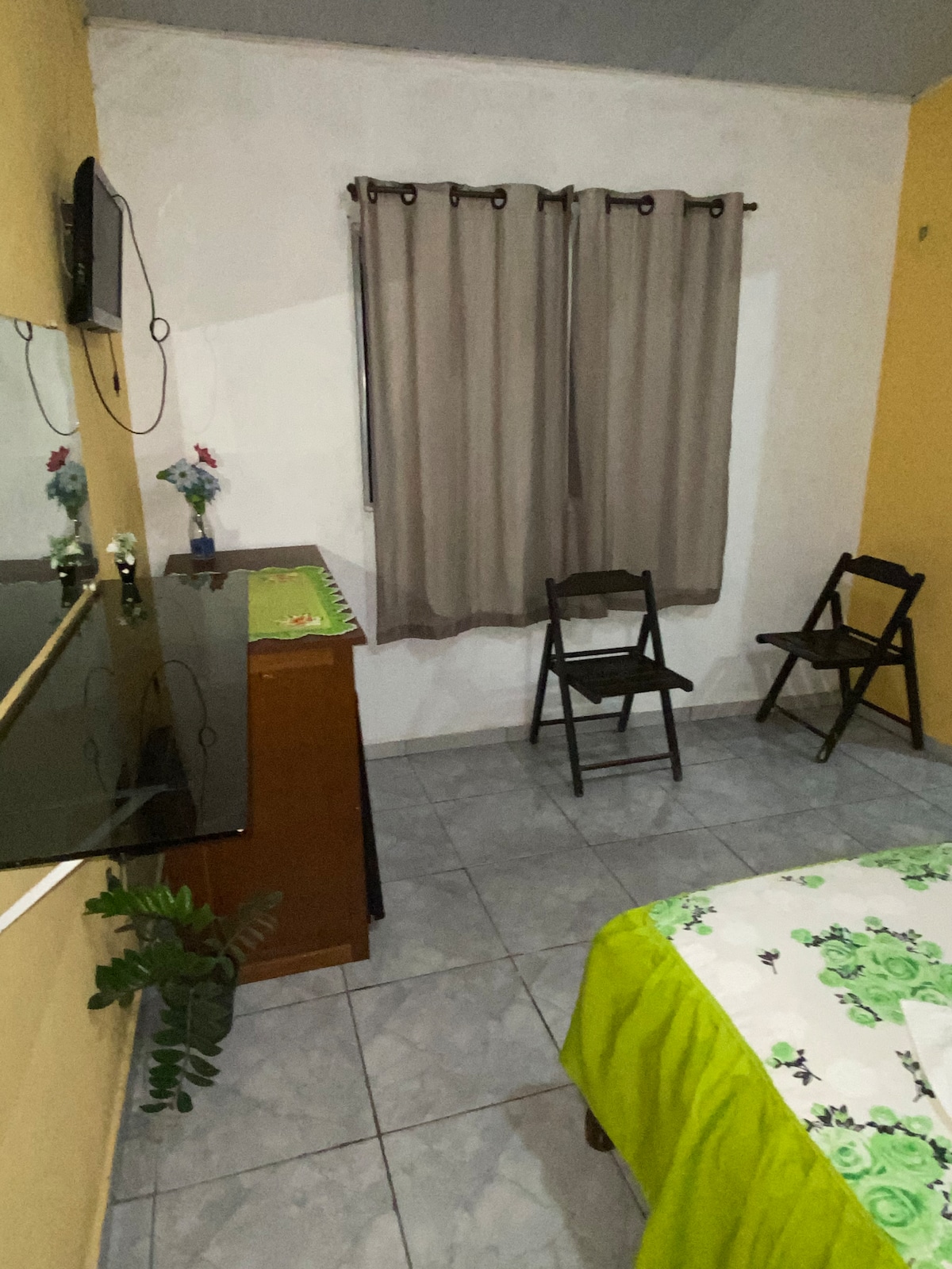 A simple sleeping area is presented with a vibrant floral bedding set resting on a spacious bed. Two black chairs are positioned next to a wooden dresser, while a small plant adds a touch of greenery. Neutral-toned walls and curtains create a calm atmosphere.