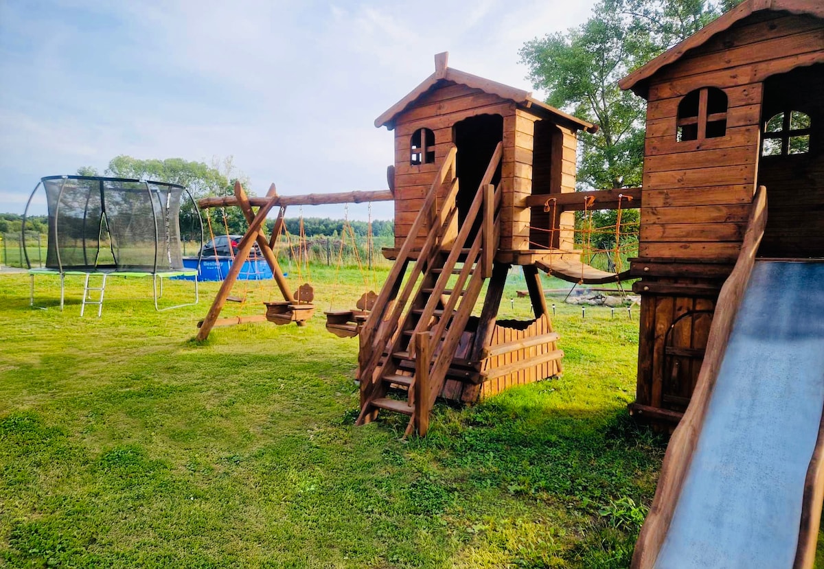 A wooden play structure is positioned on the green grass, featuring two elevated playhouses connected by a bridge and a slide. Nearby, a trampoline can be seen, and the backdrop includes trees and open fields, creating a spacious outdoor recreational area.