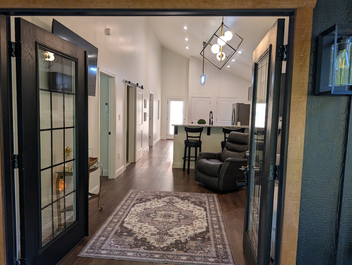 An open entryway leads into a spacious living area featuring a contemporary design. A large area rug is positioned on wood flooring. A kitchen bar with seating is visible in the background, complemented by ample natural light from the windows. Dark furnishings provide contrast.