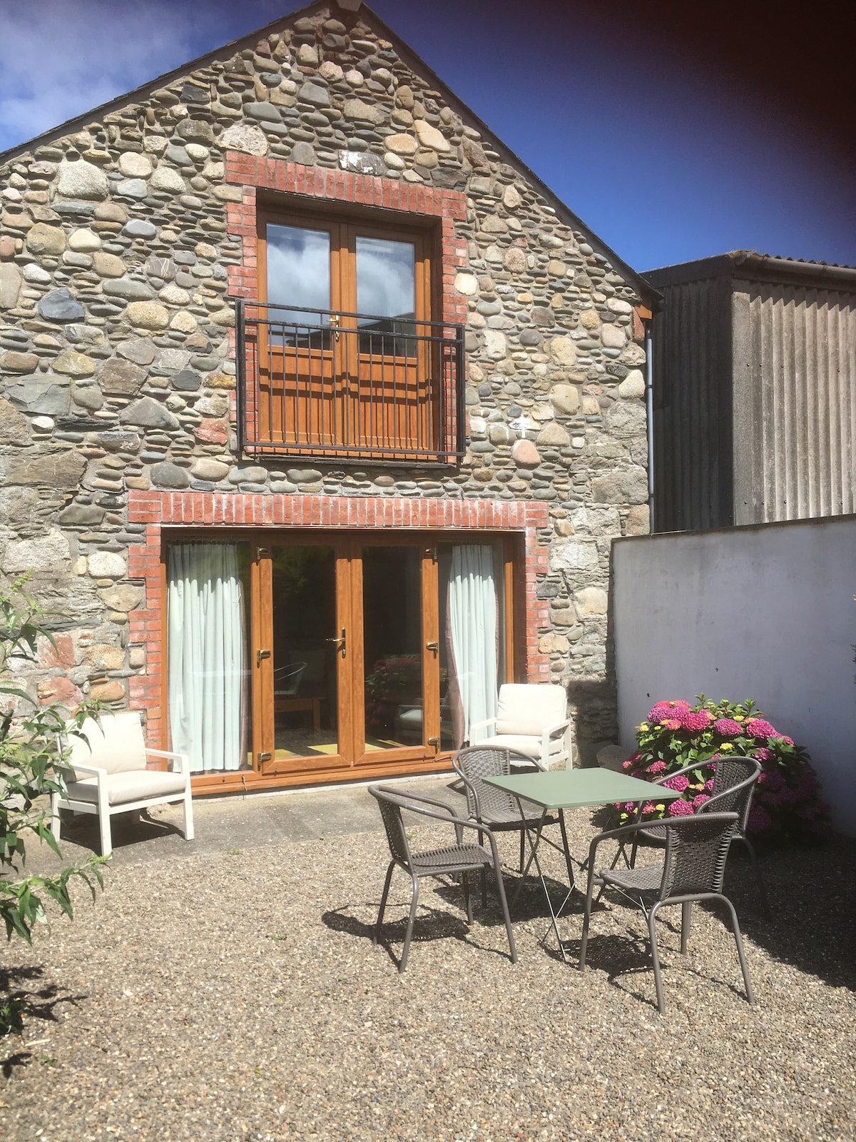 The exterior view of the cottage highlights a stone façade, complemented by wooden French doors and a balcony. A gravel garden area features a small table and four chairs, surrounded by greenery and blooming flowers, creating a tranquil outdoor space.