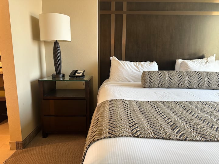 Чисто бяло спално бельо покрива леглото и нощните шкафчета от всяка страна с индивидуална лампа за удобство на гостите. 