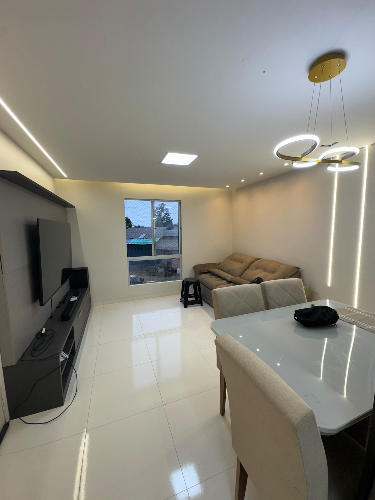 A modern living area is highlighted by a comfortable sofa beside a sleek entertainment unit. A dining table with light-colored chairs is positioned nearby, with a stylish ceiling fixture providing soft illumination. Natural light enters through a window, enhancing the bright, minimalist design.