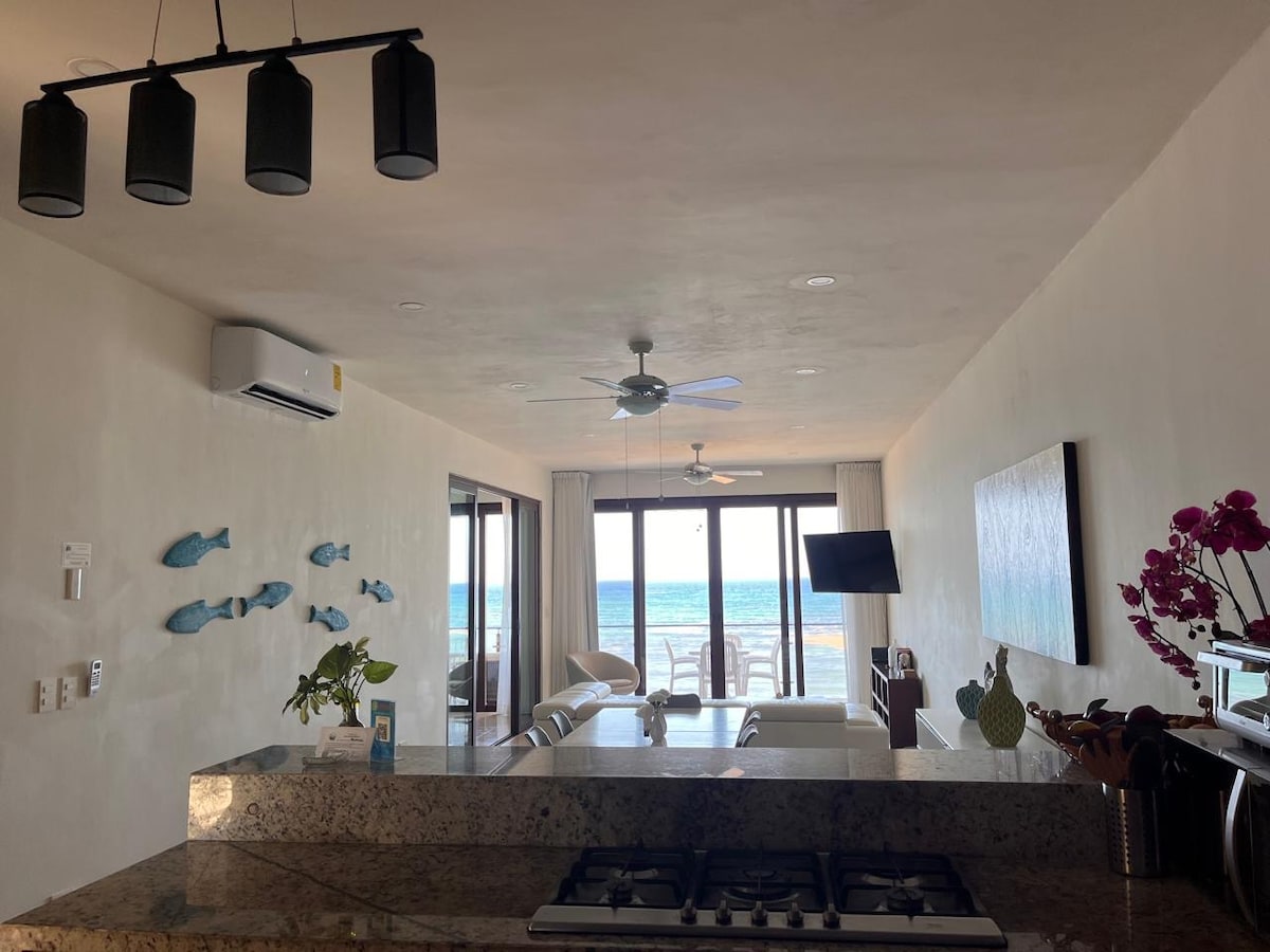 A modern living space is seen from the kitchen, featuring a sleek granite counter and decorative fish on the wall. Large glass doors open to a view of the sea, allowing natural light to fill the room. Furnishings include a comfortable sofa and a television.