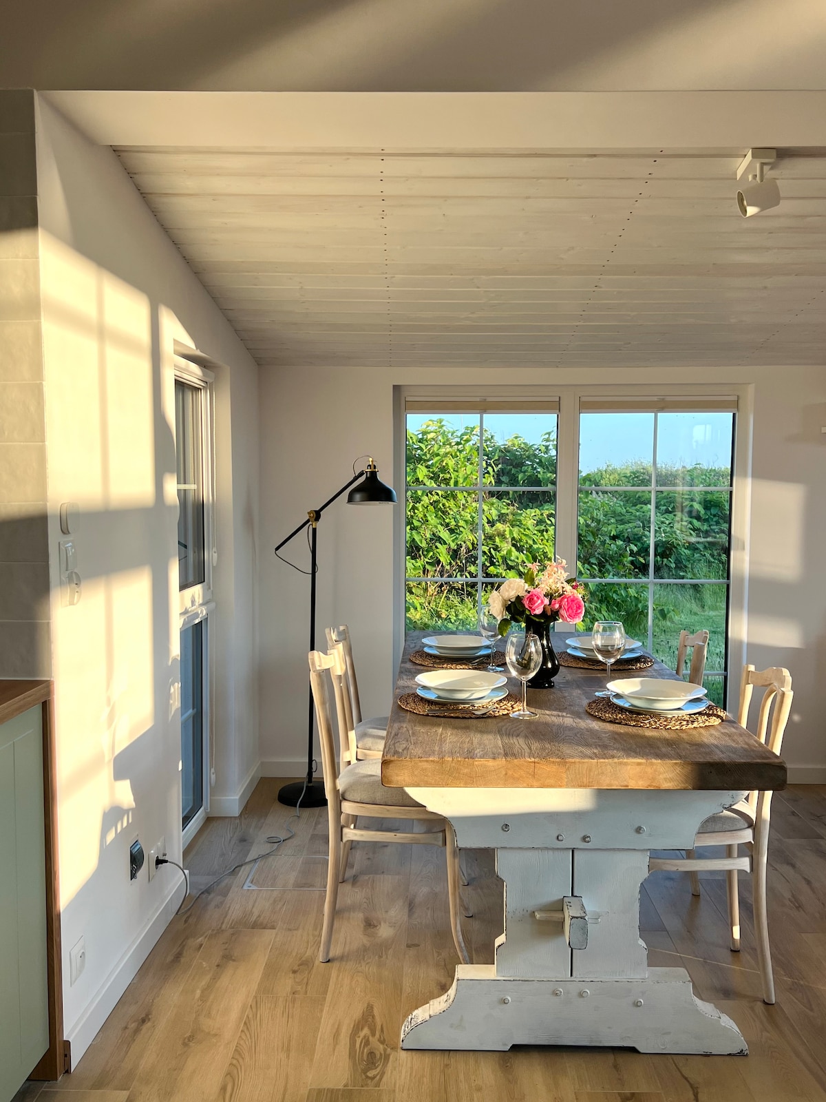 A dining area features a rustic wooden table set for a meal, complemented by white chairs. Sunlight filters through large windows, casting soft shadows on the walls. A vase with flowers and dinnerware creates a welcoming atmosphere, with greenery visible outside.
