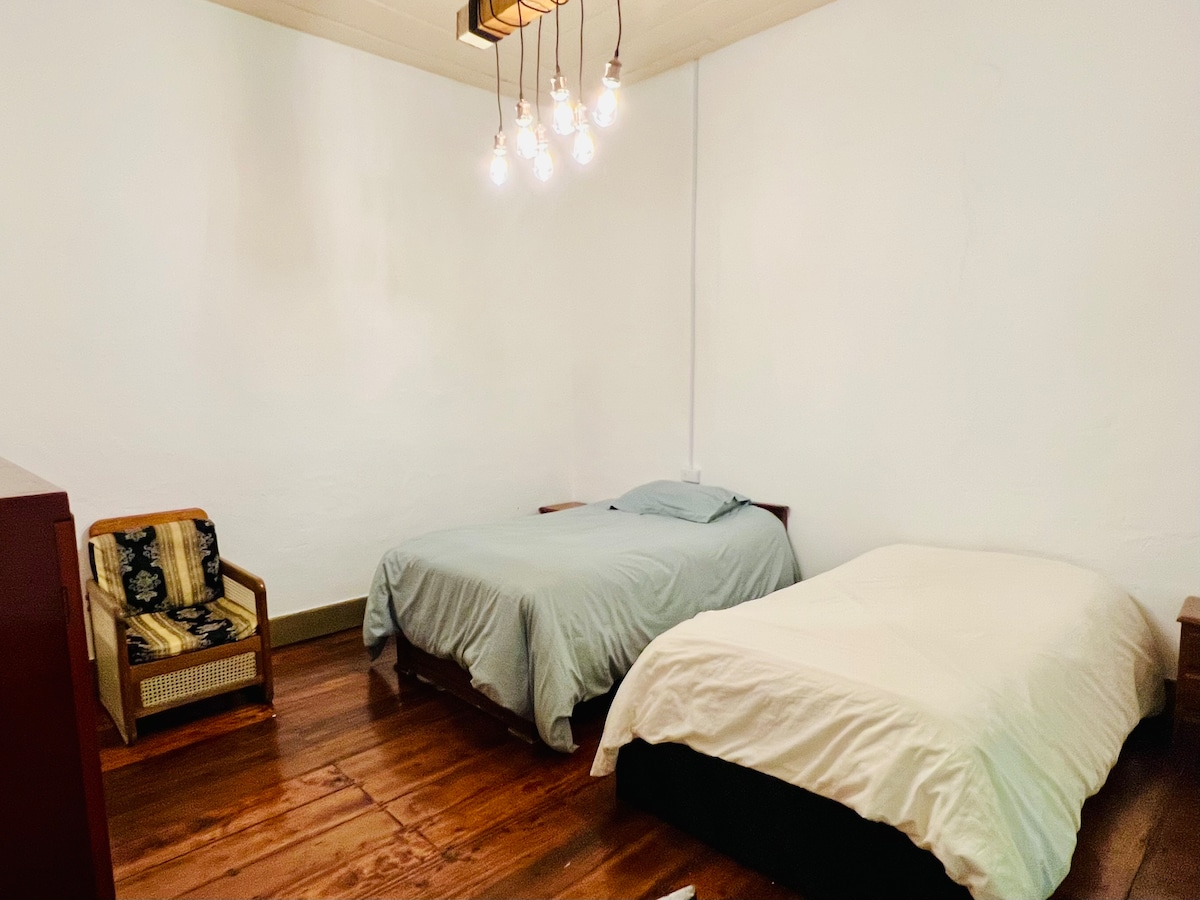 Two beds are arranged within a spacious room featuring light wooden flooring. A chair with a woven seat is placed beside the beds. A modern light fixture hangs from the ceiling, providing soft illumination, while the walls are painted in a neutral tone.