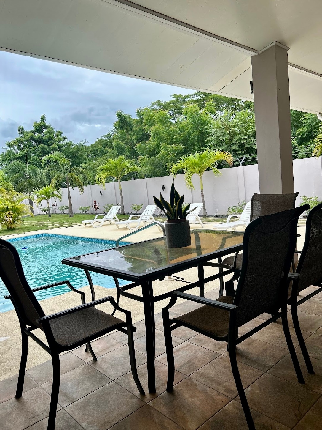 A shaded outdoor dining area includes a glass-topped table surrounded by six black metal chairs. The pool, visible in the background, is framed by lush greenery and several lounge chairs, offering a serene space for relaxation and entertaining.
