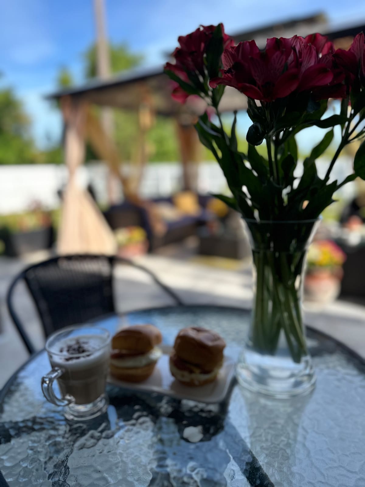 A glass-top table is set with a creamy coffee drink next to two sandwiches. A vase filled with a bouquet of flowers stands prominently, while a cozy seating area can be seen in the background, framed by lush greenery and colorful plants.