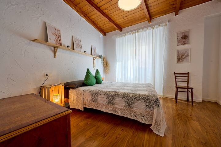 Dormitorio principal con techo de madera y vigas vistas, cama de matrimonio con colchón premium, suelo de madera y ventana con cortinas de lino. Decoración rural con cuadros botánicos y plantas