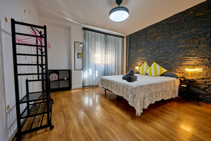 Dormitorio 2 con pared de piedra pizarra oscura, cama de matrimonio con colchón premium, suelo de madera y aire acondicionado. Diseño de contraste entre la piedra natural y la ropa de cama blanca.
