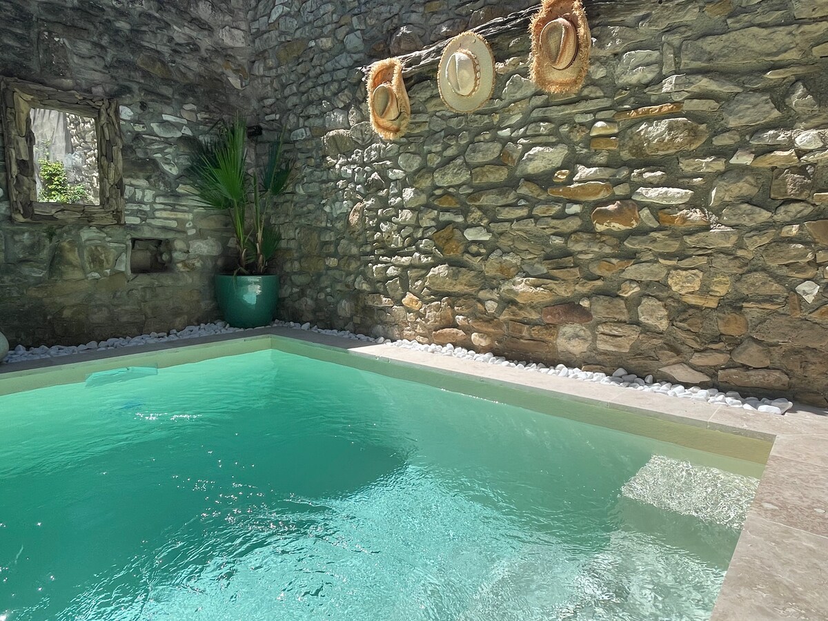 A private pool is framed by natural stone walls, creating a serene atmosphere. Three straw hats are displayed on the wall, and a vibrant green plant adds a touch of color. Light reflects gently on the clear, inviting water.