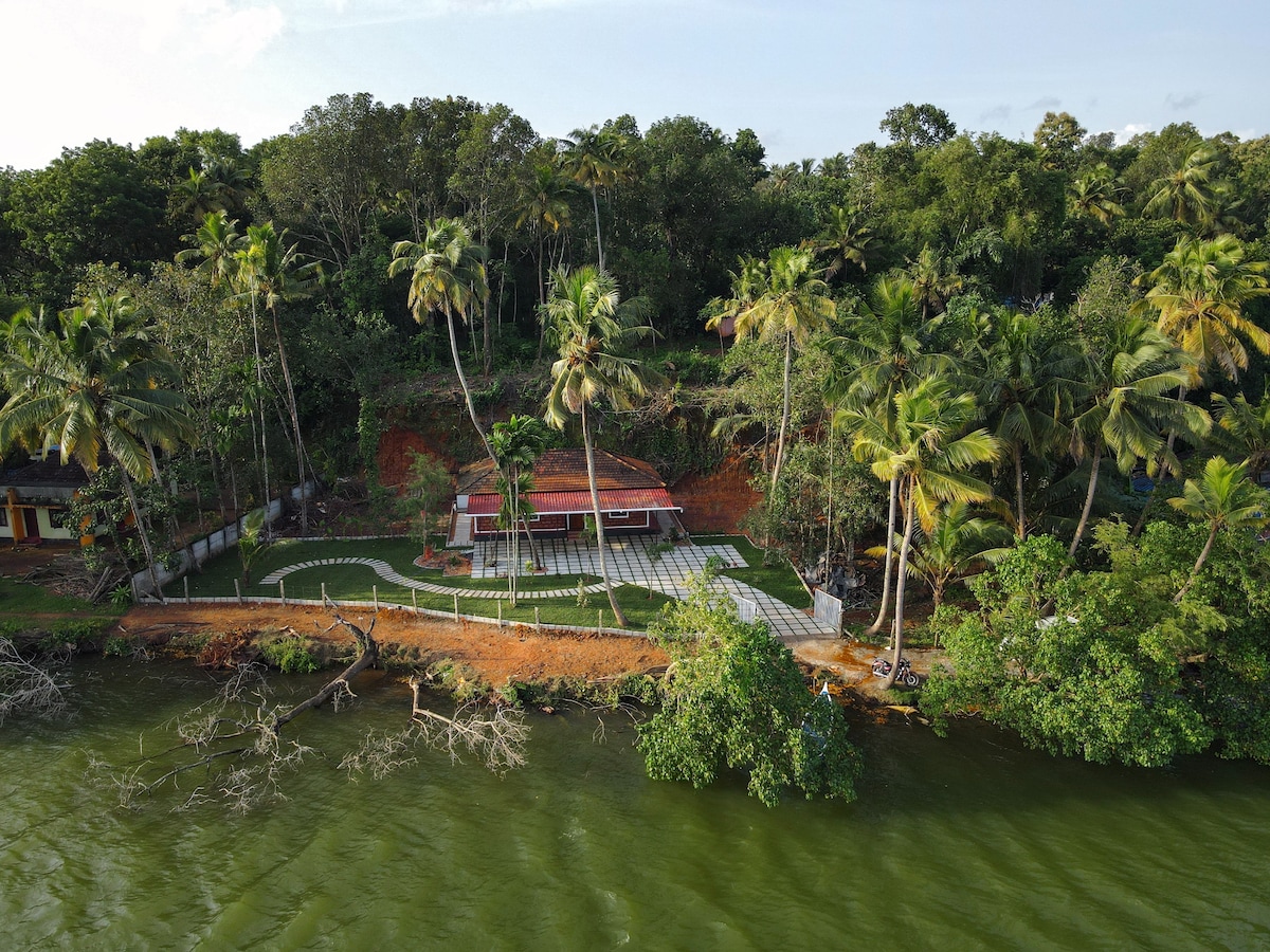 An aerial view reveals a vibrant property surrounded by lush greenery and palm trees, overlooking calm waters. A spacious patio is visible, designed for outdoor gatherings, while a red-roofed building adds a striking contrast to the natural landscape.