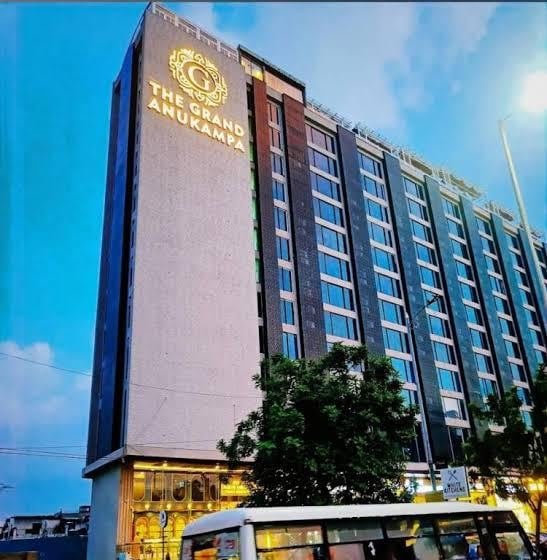 A modern hotel exterior is presented with a sleek design, featuring multiple stories and a prominent sign displaying the name 'THE GRAND ANUKAMPA.' Lush greenery surrounds the entrance, enhancing the welcoming atmosphere of the building against a backdrop of a partly cloudy sky.