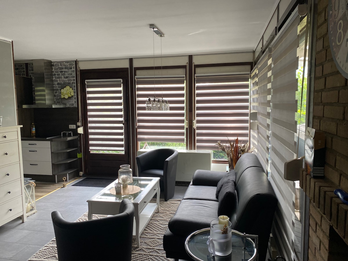 An inviting living area features modern furnishings including a black sectional sofa and a white coffee table. Large windows with dual shades allow natural light to filter in. A stylish area rug and a small side table with decorative items add to the room's comfort.